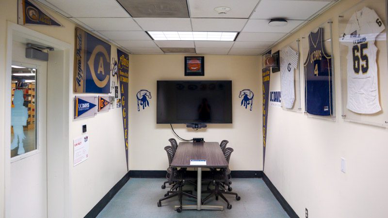 Small conference table flanked by walls decorated with Aggie pennants and framed athletic jerseys, with a mounted TV screen.