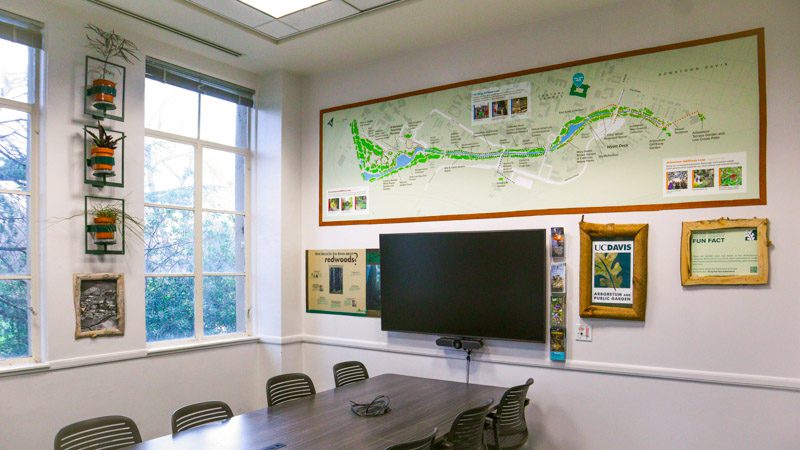 Conference table near tall windows with wall-mounted planters, a large illustrated map of the UC Davis Arboretum and nature photos above a mounted screen.