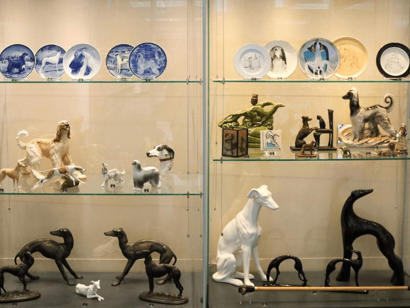 Glass case with nearly two dozen pieces of figurines, plates and other functional objects in the shape of an Afghan hound or greyhound