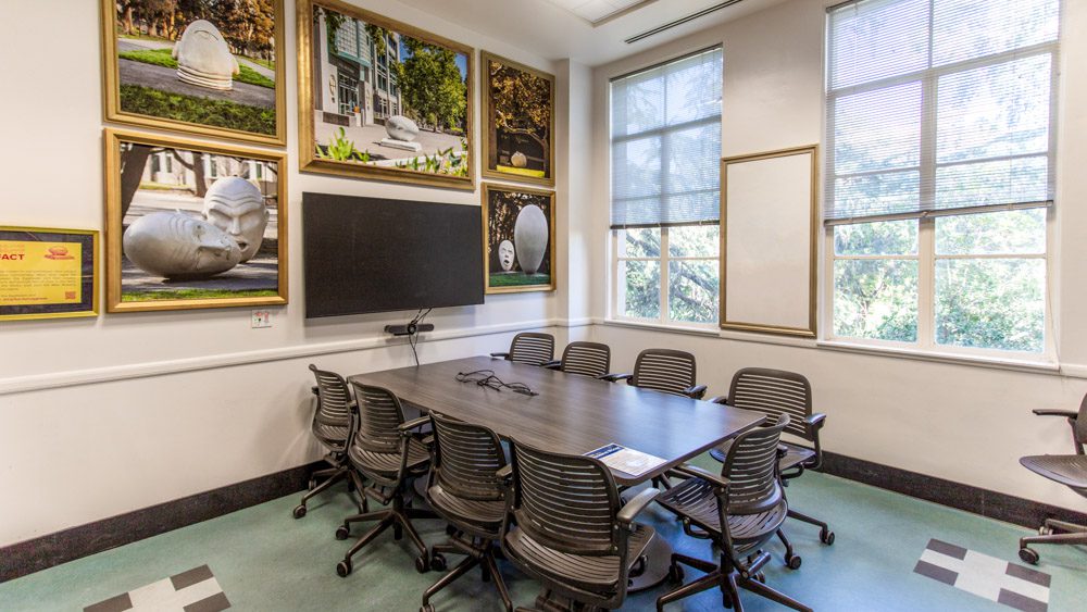 Conference table by tall windows, with a wall of framed photos of UC Davis's giant 'Egghead' sculptures and a mounted TV screen.