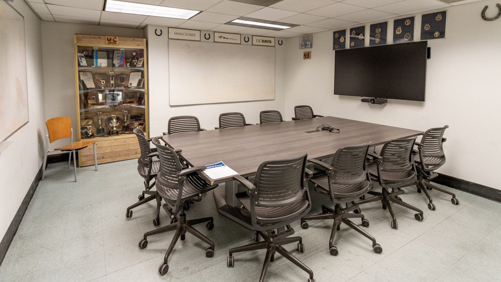 Conference table across from a rustic wood display cabinet holding trophies and awards, with a mounted TV screen and whiteboard.
