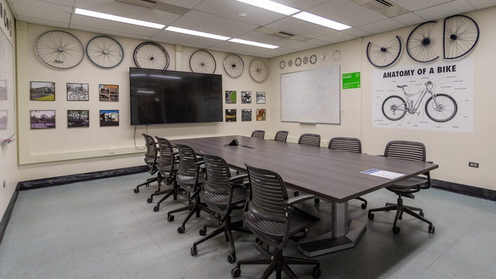 Conference table with mounted bicycle wheels lining the upper walls and an 'Anatomy of a Bike' diagram poster, with a mounted TV screen and whiteboard.