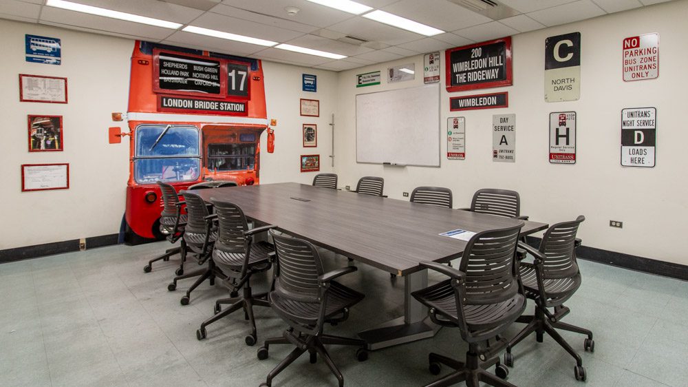 Conference table beside a wall built to resemble the front of a red Unitrans double-decker bus, with a whiteboard, transit signs and memorabilia on the walls.